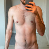 Man with nipple piercing showing torso, body hair, and beard, holding a smartphone for a selfie.