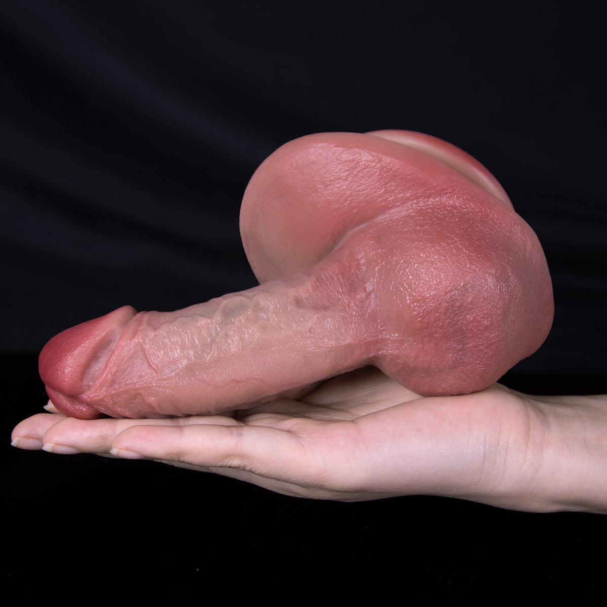 A realistic dildo is being held by a person's hand against a black background.