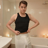 Man in black tank top and white shorts bulging noticeably in steamy bathroom.