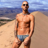 Muscular tattooed man in patterned Speedo stands on desert dunes near mountains.